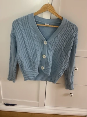 Ljusblå kabelstickad kofta - Snygg ljusblå kabelstickad kofta med stora vita knappar och v-ringning. Koftan har ribbade muddar och är något croppad i modellen 🩷