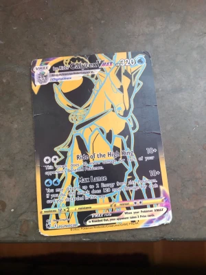 Pokémonkort Ice Rider Calyrex VMAX - Säljer ett Pokémonkort, Ice Rider Calyrex VMAX, med färgstark design i svart, blått och gult. Kortet har holografiska detaljer och är tryckt på klassiskt Pokémonkort-material. Perfekt för samlare eller dig som vill boosta din lek med en VMAX. Kortet är fejk
