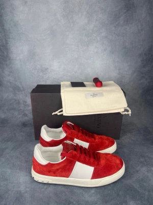 Röda och vita Valentino Garavani Flycrews - Snygga sneakers från Valentino Garavani i rött och vitt. Skorna har en låg silhuett, ovandel i mocka och läder, samt vita detaljer och nitar på sulan. Klassisk snörning och logga på insidan. Perfekta för dig som vill sticka ut med stil. Box, dustbag, extra skosnören och äkthetsbevis ingår. Storlek 42.