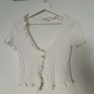 Vit ribbad blus/kofta med volangkant - Supersöt vit ribbad blus med korta ärmar, v-ringning och knappar framtill. Volangdetaljer längs halsringning, ärmslut och nederkant ger en romantisk vibe. Perfekt att styla med jeans eller kjol för en fräsch look.
