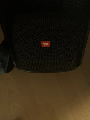 JBL svart bärbar högtalare - Stor bärbar högtalare från JBL i svart med robust plastmaterial och metallgaller framtill. Högtalaren har ett tydligt JBL-logotyp på framsidan och kontrollpanel på ovansidan. Perfekt för att ta med sig ljudet överallt.