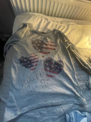 Grå långärmad t-shirt med hjärttryck - Grå långärmad t-shirt med tre hjärtan på bröstet i amerikansk flaggdesign och små glittriga stenar runt motiven. Texten 'With love from NY' står tryckt framtill. Mjuk och bekväm bomullskänsla, perfekt till jeans eller kjol.