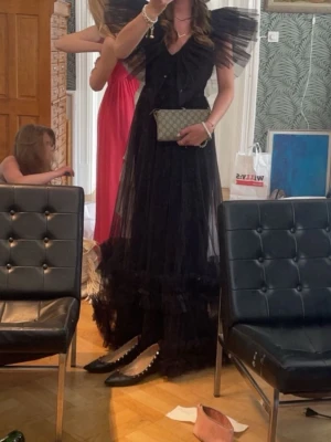 Svart balklänning med volang - Säljer en svart långklänning med dramatiska volangdetaljer på axlarna och v-ringning. Klänningen har ett lager av tunn tyll som ger en luftig och elegant känsla. Perfekt för dig som vill sticka ut på bal eller fest.