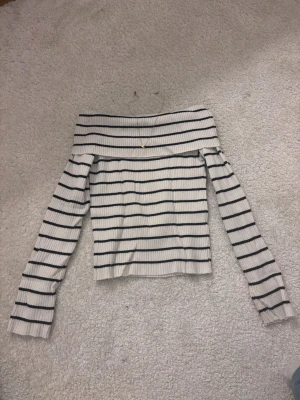Randig offshoulder topp - Säljer en vit ribbad offshoulder topp med svarta horisontella ränder. Toppen har långa ärmar och en bred kant över axlarna. Perfekt för dig som gillar en trendig och avslappnad stil.