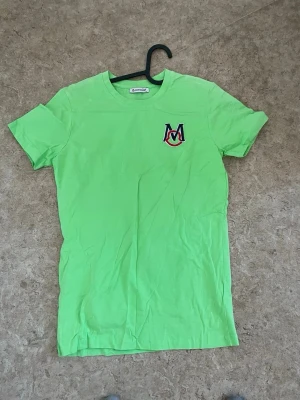 Grön t-shirt med M-logga - Snygg grön t-shirt från Mondo med en broderad M-logga i rött, svart och vitt på bröstet. Klassisk rund halsringning och korta ärmar. Tillverkad i mjuk bomull som känns skön mot huden. Perfekt för dig som gillar färg och enkel stil.