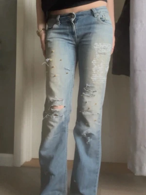 Ljusblå slitna bootcut jeans - Säljer ett par ljusblå bootcut jeans med slitningar och hål på benen. Jeansen är låg midjade och detaljerade slitningar och guld spikar framtill för en trendig look. Perfekta för dig som gillar en avslappnad och edgy stil.