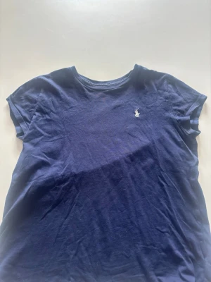 Mörkblå t-shirt från Ralph Lauren - Klassisk mörkblå t-shirt från Ralph Lauren med rund halsringning och korta ärmar. T-shirten har den ikoniska vita Polo-loggan broderad på bröstet. Tillverkad i mjuk bomull för en skön och avslappnad stil. Mycket bra skick
