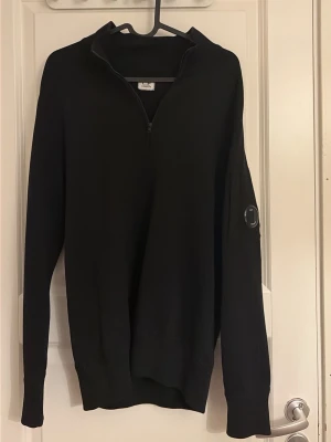 Svart half-zip tröja från C.P. Company - Svart långärmad half-zip tröja från C.P. Company med linsdetalj på ärmen. Tröjan har hög krage och ribbade muddar. Perfekt för en clean och stilren look. Pris kan diskuteras 