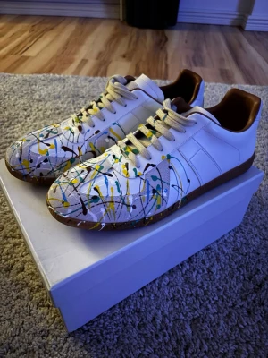 Maison Margiela sneakers med färgstänk - Unika vita sneakers från Maison Margiela med färgstänk i blått, gult och brunt. Skorna har klassisk låg siluett, vita snören och bruna detaljer på insidan samt gummisula. Tillverkade i läder och mocka för en exklusiv känsla.