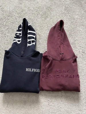 Paketpris, två hoodies från Hilfiger & Peak Performance - Säljer två snygga hoodies: en marinblå från Hilfiger med stor logga på huvan och en vinröd från Peak Performance med broderad text framtill. Båda har huva och klassisk passform, perfekta för en avslappnad streetstyle-look.