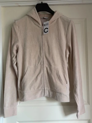 Beige velourhoodie med dragkedja - Mjuk och skön hoodie i beige velour med dragkedja framtill och två stora fickor. Klassisk huva och långa ärmar. Perfekt för en avslappnad och trendig look. Storlek M