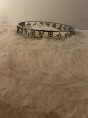 Silverfärgat armband med LOVE-text - Snyggt armband i silverfärg med utskuren texten 'LOVE' och dekorativa detaljer runt om. Armbandet har en stilren och modern design i metall och passar perfekt för dig som vill ha ett statement-smycke. Enkel att matcha med andra accessoarer.
