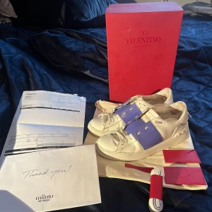 Valentino Garavani sneakers - Ett par vita och lila Valentino Garavani sneakers i skinn. Skorna har snörning och en bred lila/blå rem över mitten. De kommer med originalförpackning och tillbehör. det finns även KVITTO, köpta på miinto.