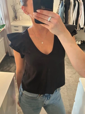 Svart topp med volangärmar - Snygg svart topp med v-ringning och volangdetaljer på ärmarna. Modellen är croppad och har en feminin siluett som passar perfekt till jeans eller kjol. 