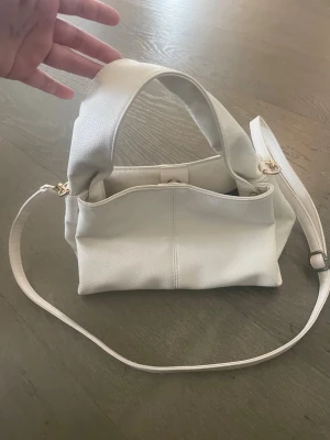 Vit axelremsväska i konstläder - Snygg vit handväska med både kort och lång axelrem. Väskan har en mjuk, lite slouchy form och är gjord i konstläder med guldfärgade metalldetaljer. Perfekt för dig som vill ha en stilren och enkel accessoar till din outfit.