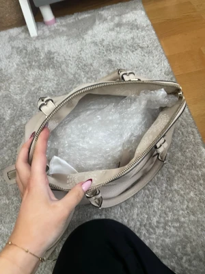 Beige handväska i äkta läder med detaljer.( kom med prisförslag!!) - Snygg beige handväska i mjukt, strukturerat läder med guldfärgade metalldetaljer och dragkedja. Väskan har både handtag och axelrem samt en praktisk ytterficka med dragkedja. Perfekt för dig som vill ha plats för allt du behöver och gillar stilrena accessoarer.