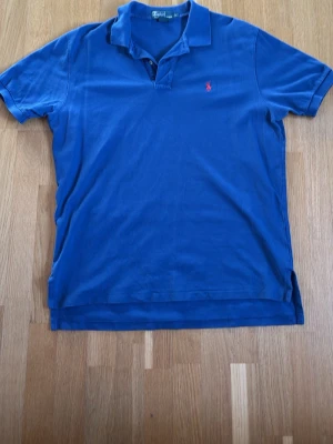 Blå pikétröja från Ralph Lauren - Klassisk blå pikétröja från Ralph Lauren med röd broderad logga på bröstet. Tröjan har korta ärmar, krage och knappar vid halsen. Tillverkad i mjuk bomull som känns skön mot huden. Perfekt för en avslappnad och stilren look. Storlek L och sitter bra på en kille som är 190+.