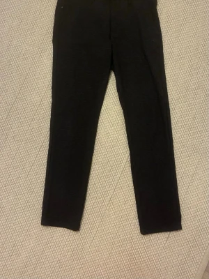 Svarta smala kostymbyxor - Säljer ett par svarta kostymbyxor med smal passform och diskret struktur i tyget. Byxorna har raka ben och två bakfickor, perfekta för en clean och stilren look. Inget synligt märke men materialet känns mjukt och stretchigt.