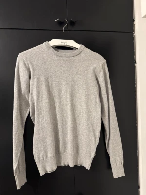 Ljusgrå stickad tröja - En stilren ljusgrå stickad tröja med rund halsringning och långa ärmar. Tröjan har ribbade muddar vid ärmslut och nederkant, vilket ger en klassisk och enkel look. Perfekt att matcha med jeans eller kjol för en avslappnad stil.