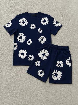 Mörkblått Denim Tears set - Snyggt tvådelat set med t-shirt och shorts i mörkblått, dekorerat med stora vita blommönster över hela plaggen. Setet har en avslappnad passform och elastisk midja på shortsen. Perfekt för en chill och trendig look. Tröjan är M och shortsen är M/L( pris kan diskuteras)