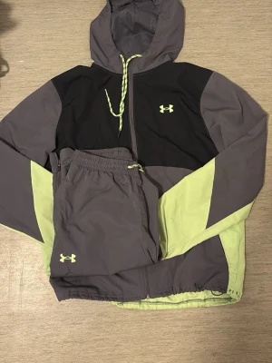 Under Armour träningsset grå/gul - Snyggt träningsset från Under Armour med jacka och byxor i grått och ljusgult. Jackan har huva, dragkedja och snörning, medan byxorna har resår i midjan och snörning. Båda plaggen har Under Armour-logga i gult. Perfekt för träning eller chill.