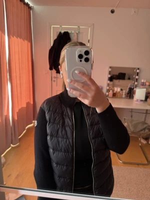 Svart dunjacka från Moncler - Snygg svart dunväst från Moncler med quiltad design och hög krage. Västen har dragkedja framtill och Moncler-logga på ena axeln. Perfekt att slänga över en hoodie eller tröja för en trendig lager-på-lager-look.
