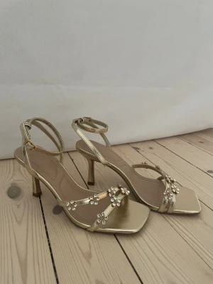Guldiga klackskor med blommor  - Helt oanvända guldiga sandaletter med smala remmar och dekorativa blommor med strassdetaljer. Skorna har fyrkantig tå, ankelrem med spänne och en elegant klack på 8 cm.