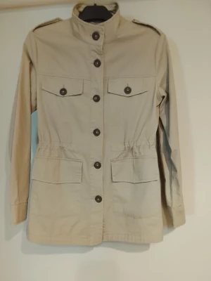 Beige rock med ståkrage och fickor - Stilren beige rock med ståkrage och stora knappar framtill. Fyra fickor med lock och knapp, samt axelklaffar för en cool militärinspirerad look. Tillverkad i ett lätt bomullstyg, perfekt för vår, sommar och höst. Ny.