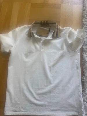 Vit Burberry skjorta  - Snygg vit skjorta från Burberry med klassisk rutigt mönster på kragen. Skjortan har korta ärmar och är tillverkad i mjuk bomull. Perfekt för en stilren och trendig look.