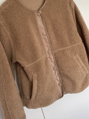Beige teddyjacka från H&M XS - Supermjuk teddyjacka i beige från H&M. Jackan har rund hals, tryckknappar framtill och två fickor. Perfekt för lager-på-lager och ger en mysig vibe. Tillverkad i fluffigt teddy-material som håller dig varm och trendig.