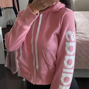 Rosa Adidas hoodie med dragkedja - Säljer en rosa hoodie från Adidas med vit dragkedja och vita snören i huvan. Stor Adidas-logga tryckt längs ena ärmen. Perfekt för en sportig och avslappnad stil. Mjuk bomullsblandning och två fickor framtill.