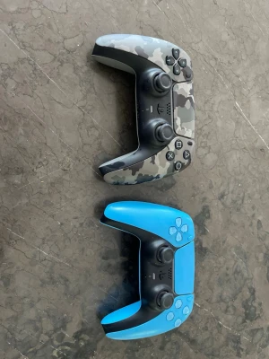 PS5 DualSense handkontroller  - Två PlayStation 5 DualSense handkontroller, en i grått camouflagemönster och en i klarblå färg. Båda har svart mittparti och klassiska PlayStation-knappar. Ergonomisk design i plast, perfekt för gaming på PS5. Typ oanvänd. 250 kr för en 450 för båda 