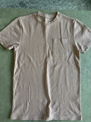 Beige ribbad t-shirt med bröstficka - Snygg beige t-shirt med diskret ribbad struktur och en praktisk bröstficka. Klassisk rund halsringning och korta ärmar. Perfekt för en avslappnad och stilren look.