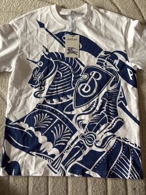 Vit Burberry t-shirt med blått tryck - Snygg vit t-shirt från Burberry med stort blått tryck av riddare och häst på framsidan. Klassisk rund halsringning och korta ärmar. Perfekt statement-plagg för dig som gillar exklusiva märken och unik design.