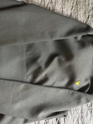 Svart hoodie från Lyle & Scott - Svart hoodie från Lyle & Scott. Klassisk design med gul logotyp på bröstet och stor magficka. Mjukt material och ribbade muddar vid ärmsluten. Använd ett fåtal gånger.