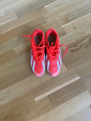 Röda Adidas fotbollsskor med vita ränder - Säljer ett par snygga röda Adidas fotbollsskor med klassiska vita ränder på sidorna. Skorna har snörning och en yttersula med gula detaljer och dobbar för bra grepp på planen. Perfekta för dig som vill sticka ut på fotbollsplanen.