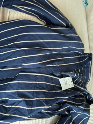 Blå randig skjorta från H&M - Snygg blå skjorta med vita vertikala ränder från zara Skjortan har klassisk krage, knäppning framtill och långa ärmar. Perfekt för en stilren och trendig look. Tillverkad i mjuk bomull som känns skön mot huden.