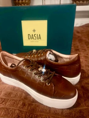 Bruna sneakers i skinn från Dasia - Snygga bruna sneakers från Dasia i croco-präglat skinn med vita tjocka sulor. Skorna har guldfärgade öljetter, bruna skosnören och en liten gulddetalj vid snörningen. Perfekta för dig som gillar en trendig och stilren look. Nyborgs i butik 1299kr 