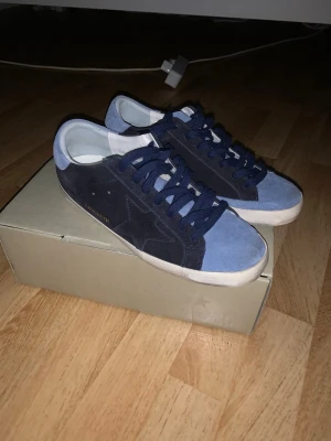 Blåa suede sneakers från Golden Goose - Snygga sneakers från Golden Goose i blå mocka med mörkblå detaljer och vita sulor. Klassisk låg modell med stjärndetalj på sidan och matchande blå skosnören. Perfekta för dig som gillar stilrena och trendiga skor.