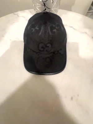 Svart keps från Gucci med GG-mönster - Snygg svart keps från Gucci med klassiskt GG-monogram över hela kepsen. Kepsen har böjd skärm och är tillverkad i ett tygmaterial med läderdetaljer längs skärmen. Perfekt accessoar för en trendig streetstyle.