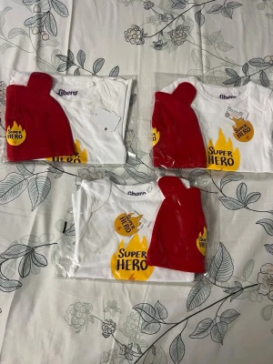 Super Hero bodyset från Libero - Säljer ett set med vit body och röd cape från Libero. Bodyn har trycket 'Super Hero' i gult och svart framtill och är i mjuk bomull. Perfekt för små superhjältar! Capen är röd och fästs enkelt på bodyn.