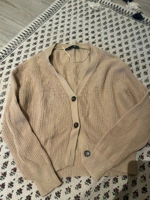 Beige stickad kofta från 157 - Beige stickad kofta från 157 med v-ringning och tre knappar framtill. Koftan har ribbstickade muddar och är tillverkad i ett mjukt, grovstickat material. Perfekt att slänga över en t-shirt eller linne för en chill look.