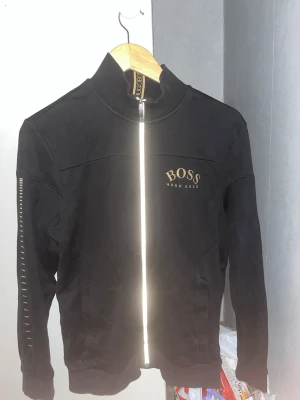 Svart ziptröja från Hugo Boss  - Snygg svart ziptröja från Hugo Boss med guldfärgad logga på bröstet och detaljer på ärmen. Tröjan har hög krage, dragkedja framtill och ribbade muddar. Perfekt för en stilren och sportig look. Kan sälja silver halsband och tröjan för 1200kr
