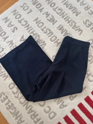 Vida svarta kostymbyxor - Snygga svarta kostymbyxor med vida ben och hög midja. Byxorna har pressveck framtill och stängs med dragkedja och knapp. Perfekta för en stilren och trendig look. Materialet känns mjukt och följsamt.