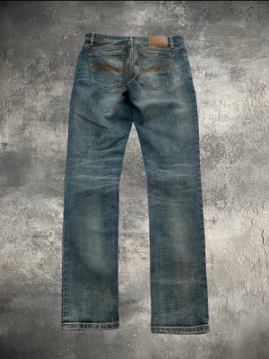 Blå Spodnie Skinny Lin Nudie Jeans - Säljer ett par klassiska blå jeans från Nudie Jeans med raka ben och femficksmodell. Jeansen har snygga slitningar och kontrastsömmar, samt läderpatch bak i midjan. Perfekta för en avslappnad och trendig look. Pris kan diskuteras.