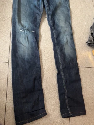 Mörkblå jeans med nitar - Snygga mörkblå jeans med slitning på låret och coola bruna nitar på bakfickan. Jeansen har en rak passform och klassiska sömmar. Perfekta för dig som gillar detaljer och en lite tuffare stil.