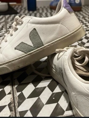 Vita Veja sneakers med lila detalj - Snygga vita Veja sneakers i lågt snitt med grå logga på sidan och lila detalj på hälen. Skorna har vita snören och ovandel i läder. Perfekta för dig som gillar stilrena och trendiga sneakers med en twist. Storleken är lite liten, säljaren som sålde dem till mig ska få köpa dem igen 