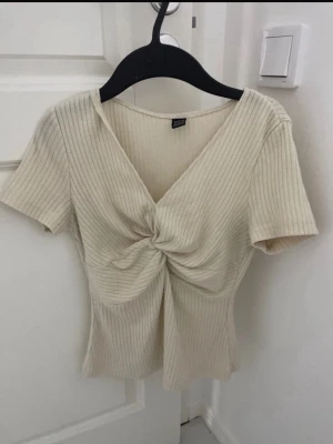 Ribbad beige topp med twist framtill - Trendig ribbad topp från Gina Tricot i beige med v-ringning och snygg twistdetalj framtill. Kortärmad och figurnära passform, perfekt till jeans eller kjol. Enkel men stilren design som passar till många looks.