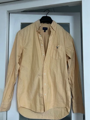 Beige skjorta från Gant - Klassisk beige skjorta från Gant med lång ärm och bröstficka med broderad logga. Skjortan har knappar framtill och är tillverkad i mjuk bomull. Perfekt för en stilren och avslappnad look.