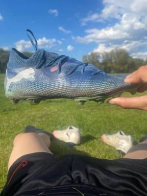 Blåa Puma fotbollsskor med dobbar - Säljer ett par blåa fotbollsskor från Puma med vit logga på sidan. Skorna har låg profil, snörning och slitstark ovandel i syntetmaterial. Dobbarna är gröna och anpassade för spel på gräs. Perfekta för dig som vill ha bra grepp och kontroll på planen.
